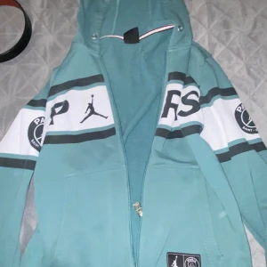 Turkos hoodie med dragkedja från Jordan - Säljer en snygg turkos hoodie från Jordan med dragkedja. Den har en cool design med svarta och vita detaljer samt en stor logga på ärmarna. Perfekt för vardagsbruk eller sportiga aktiviteter. Huvan är justerbar och det finns praktiska fickor framtill.