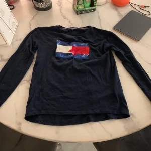 Mörkblå  långärmad tröja med paljettdetalj - Säljer en mörkblå långärmad tröja från Tommy Hilfiger med en cool paljettdetalj i form av en flagga på bröstet. Tröjan har en liten logga på ärmen och är perfekt för en avslappnad stil.