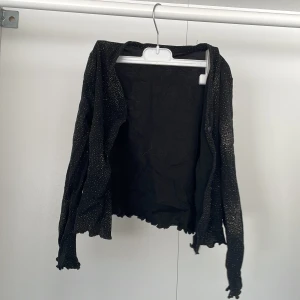 Svart glittrig kofta från Occasions - Säljer en svart glittrig kofta från Occasions, perfekt för att lägga till lite glamour till din outfit. Koftan har långa ärmar och är tillverkad i 100% polyamid. Passar för barn i åldern 8 år eller med längd 128 cm.