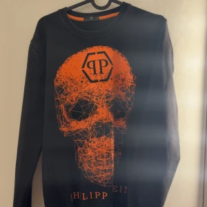 Svart tröja med orange dödskalle från Philipp Plein - Säljer en svart tröja från Philipp Plein med en stor orange dödskalle på framsidan. Tröjan har långa ärmar och en rund halsringning. Perfekt för dig som gillar en edgy stil!. Liten i storlek så skulle säga att den är mer S