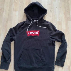 Svart hoodie från Levi's - Säljer en svart hoodie från Levi's med det klassiska röda logotyptrycket på bröstet. Tröjan har en stor ficka framtill och justerbar huva med vita snören. Perfekt för en avslappnad stil.