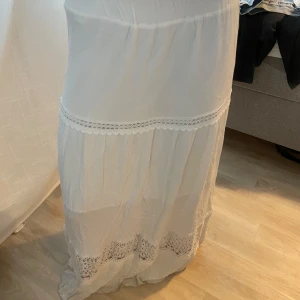 Vit långkjol med spetsdetaljer - Säljer en elegant vit långkjol med vackra spetsdetaljer. Kjolen har en luftig och flödande design som ger en romantisk känsla. Perfekt för varma dagar eller speciella tillfällen. Det står ingen storlek men gissar på xs/s. Liten defekt förekommer längst ned