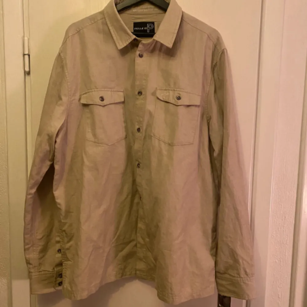 Säljer en stilren beige skjorta från Pelle P i storlek XXL. Skjortan har långa ärmar och två bröstfickor med knappar. Perfekt för en avslappnad men ändå snygg look.. Paidat.