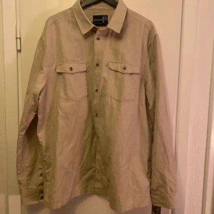Beige skjorta från Pelle P - Säljer en stilren beige skjorta från Pelle P i storlek XXL. Skjortan har långa ärmar och två bröstfickor med knappar. Perfekt för en avslappnad men ändå snygg look.