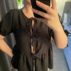 Svart och vit blus med knytning - Säljer två blusar i svart och vit färg med knytning framtill. Blusarna har korta ärmar och en volangdetalj nedtill. Perfekta för en stilren look . Säljer båda två om man vill ha en utav de så är de bara o skriva 