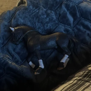 Svart leksakshäst - En svart leksakshäst med vita detaljer på benen och en vit fläck på huvudet. Hästen har en realistisk design med flätad man och svans. Perfekt för samlare eller som leksak för barn. Tränset ingår (du kan få sadel med) 