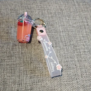 Nyckelring med rosa dryck och rem - Charmig nyckelring med en rosa dryck i en liten plastmugg med lock och sugrör. Muggen har en jordgubbsdetalj och glittrig vätska. Fäst vid en rosa rem med texten 'Happy Moments' och små blommor.