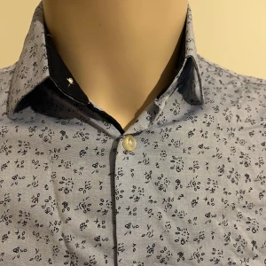 Blommig skjorta från Selected Homme - Snygg blommig skjorta från Selected Homme i en ljusblå nyans med mörkare blommönster. Skjortan har en slim fit passform och är långärmad med knappar framtill. Perfekt för en stilren look!