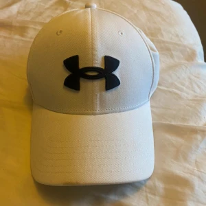 Vit keps från Under Armour - Snygg vit keps från Under Armour med svart logga framtill. Perfekt för sportiga aktiviteter eller som en stilren accessoar. Kepsen har en klassisk design med böjd skärm och justerbar passform.