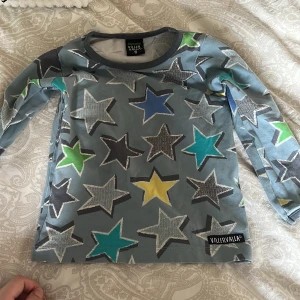 Stjärnmönstrad långärmad tröja från Villervalla - Säljer en långärmad tröja från Villervalla i ekologisk bomull. Tröjan har ett coolt stjärnmönster i blått, grönt, gult och svart på en grå bas. Perfekt för lekfulla dagar!