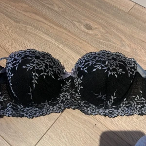 Svart spets-bh med rosett - Elegant svart bh med spetsdetaljer och en söt rosett framtill. Den har en klassisk design med justerbara axelband och hakknäppning bak. Perfekt för en stilfull look.