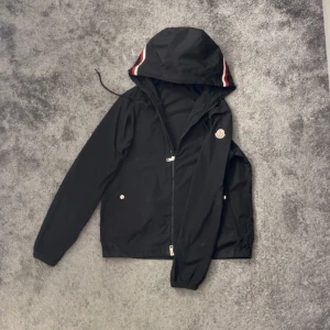 Svart Moncler Windbreaker - Säljer en stilren svart vindjacka från Moncler med huva och dragkedja. Jackan har vita, blåa och röda detaljer på huvan och två fickor med knappar framtill. Perfekt för blåsiga dagar. Äkthet kan bevisas skicka meddelande privat så kan jag visa.