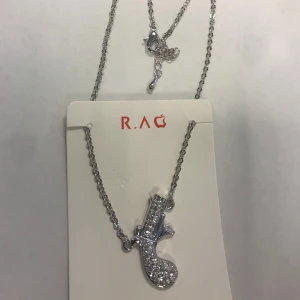 Strass Silverfärgat halsband med glittrig pistolberlock - Snyggt silverfärgat halsband från R.A.G med en unik pistolformad berlock täckt av glittrande stenar. Perfekt för att ge en edgy touch till din outfit. Kedjan är tunn och elegant, vilket gör den lätt att bära.  Helt ny 