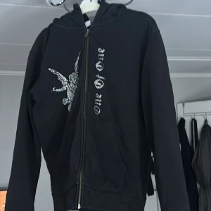 Svart hoodie med dragkedja från One of One - Säljer en svart hoodie med dragkedja från One of One. Den har ett unikt tryck på framsidan och en bekväm huva. Perfekt för en avslappnad stil. Nypris=1500