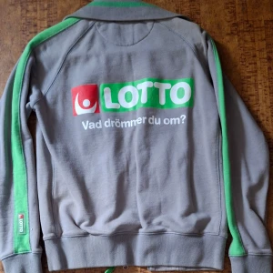 Grå Lotto jacka med gröna detaljer - Säljer en grå jacka från Sv. Spel.   Lotto med gröna ränder längs ärmarna och dragkedja framtill. Jackan har texten 'Vad drömmer du om?' tryckt på ryggen. Perfekt för en sportig look!