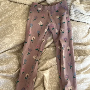 Blommiga leggings från Lindex - Säljer ett par söta leggings från Lindex i storlek 110. De är lila med ett blommigt mönster i olika färger. Perfekta för barn som gillar färgglada kläder.