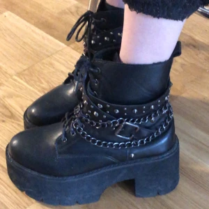 Svarta boots med kedjor och nitar - Snygga svarta boots med hög platåsula och klack. Skorna har snörning och är dekorerade med kedjor och nitar för en tuff look. Perfekta för att ge din outfit en edgy touch.
