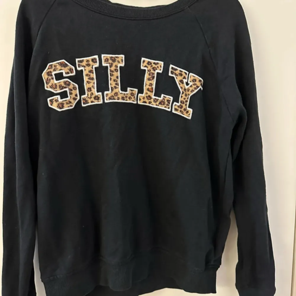 Säljer en svart sweatshirt från FB Sister med texten 'SILLY' i leopardmönster på framsidan. Tröjan har långa ärmar och en rund halsringning. Perfekt för en avslappnad stil.. Neuletakit & Villapaidat.