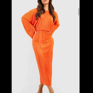 Orange långklänning - Snygg orange långklänning med plisserad design och långa ärmar. Perfekt för en färgstark look. Klänningen har en lös passform som ger en bekväm och trendig stil.