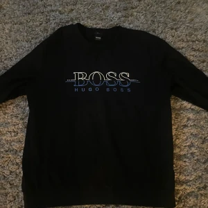 Svart tröja från Hugo Boss - Snygg svart tröja från Hugo Boss med deras ikoniska logga i vitt och blått på bröstet. Tröjan har långa ärmar och en klassisk passform. Perfekt för en stilren look.