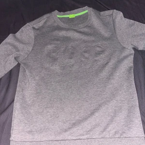 Grå tröja från BOSS - Säljer en stilren grå tröja från BOSS med subtilt präglad logotyp på framsidan. Tröjan har långa ärmar och en bekväm passform, perfekt för en avslappnad look. Den är tillverkad i ett mjukt material som känns skönt mot huden.