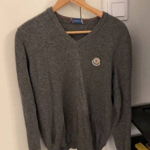 Grå tröja från Moncler - Säljer en stilren grå tröja från Moncler med v-ringning och broderad logga på bröstet. Tröjan har långa ärmar och ribbade muddar vid ärmslut och nederkant. Perfekt för kyligare dagar.