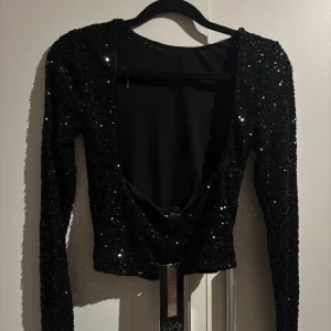 Svart glittrig festtopp - Snygg svart glittrig topp med långa ärmar och öppen rygg. Aldrig använd med prislappen kvar. Storlek XS