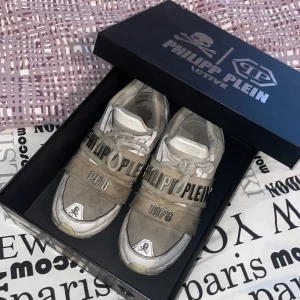 Sneakers från Philipp Plein - Snygga sneakers från Philipp Plein i beige och vit med märkets logga och årtalet 1978 på sidan. Skorna har snörning och en cool dödskalle-detalj framtill. Perfekta för en stilren och modern look.