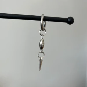 Silverfärgat örhänge i rostfritt stål  - Stilrent silverfärgat örhänge med en hängande nit. Örhänget har en enkel ringdesign som fäster i örat och en avlång pärla som leder ner till niten Perfekt för att ge en edgy touch till din stil.