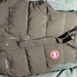 Svart dunväst från Canada Goose - Säljer en svart dunväst från Canada Goose med ett broderat märke på bröstet. Västen har dragkedja och knappar framtill samt praktiska fickor. Perfekt för kyliga dagar.