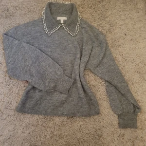 Grå stickad tröja med pärlkrage - Säljer en elegant grå stickad tröja med en unik pärlkrage. Tröjan har långa ärmar och är perfekt för kyligare dagar. Den ribbade strukturen ger en skön passform och stil. 