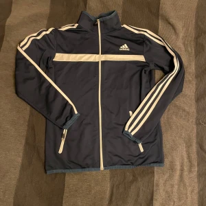 Mörkblå träningsjacka från Adidas - Säljer en mörkblå träningsjacka från Adidas med vita detaljer och dragkedja. Jackan har långa ärmar med de klassiska tre ränderna längs ärmarna och en hög krage. Perfekt för sportiga aktiviteter.