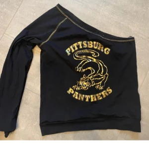 Svart offshoulder tröja med gult tryck - Säljer en cool svart offshoulder tröja med gult tryck av en panter och texten 'Pittsburg Panthers'. Tröjan har en unik design med en axel bar och långa ärmar. Perfekt för en avslappnad stil.