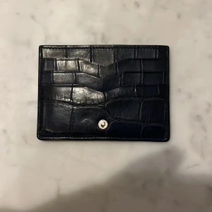 Montblanc plånbok i crocodil läder  - Snygg svart plånbok i skinn med krokodilpräglat mönster. Den har en knappdetalj på framsidan och flera fack för kort och sedlar. Perfekt för att hålla ordning på dina värdesaker med stil.