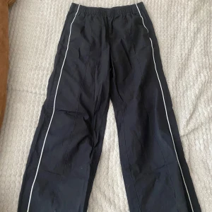 Svarta byxor från H&M - Säljer ett par svarta byxor från H&M med elastisk midja och vita ränder längs sidorna. Perfekta för en avslappnad stil.