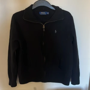 Svart tröja från Polo Ralph Lauren - Säljer en stilren svart tröja från Polo Ralph Lauren med dragkedja och broderad logga på bröstet. Tröjan har långa ärmar och är gjord av ull. Storlek är S och sitter ganska litet skulle säga att den är ner mot XS.
