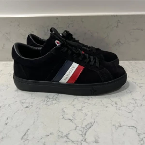 Moncler Skor - Snygga svarta sneakers från Moncler i mocka med klassiska ränder i rött, vitt och blått på sidan. Skorna har snörning och en diskret logga på plösen. Perfekta för en stilren look.