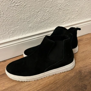Svarta boots i mocka - Snygga svarta boots i mocka med vit sula storlek 40. Använd några få gånger men har fått en skada på insidan av ena skon, se bild två, annars i fint skick. Skriv för fler bilder eller mått!