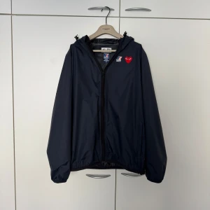 K-Way x Comme des Garçons - Säljer en stilren navy vindjacka från K-Way i samarbete med Comme des Garçons. Jackan har en dragkedja framtill och ett rött hjärta som detalj på bröstet. Perfekt för blåsiga dagar med sin lätta och funktionella design. Priset är inte hugget i sten 🪨