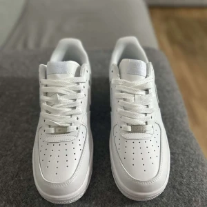 Vita Nike Air Force sneakers - Säljer ett par klassiska vita Nike Air Force sneakers. De har en stilren design med perforerade detaljer på tån och en bekväm sula. Perfekta för en casual look.
