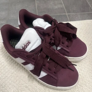 Vinröda sneakers från Adidas - Snygga vinröda sneakers från Adidas med vita detaljer och klassiska tre ränder på sidan. Skorna är i väldigt bra skick. Passar mig som är en normal 39 