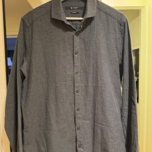 Grå skjorta från Tiger of Sweden - Snygg grå skjorta från Tiger of Sweden med diskret mönster. Skjortan har en slim passform och långa ärmar, perfekt för en stilren look. Knappar framtill ger en klassisk touch till sommar/vinter! Skick 9/10, storlek L