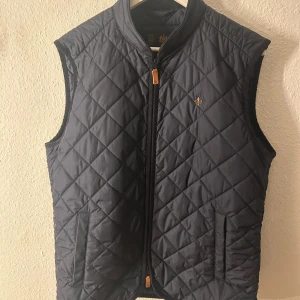 Mörkblå quiltad Morris väst - Snygg mörkblå quiltad väst med en broderad detalj på bröstet. Perfekt för lager-på-lager under kyligare dagar eller varmare dagar . Västen har en stilren design och är knappt använd. Skick 9/10