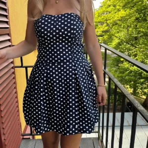 Svart prickig klänning - Charmig svart klänning med vita prickar och axelbandslös design. Perfekt för en somrig look. Klänningen har en dragkedja i sidan för enkel påklädning och en söt, utsvängd kjol.