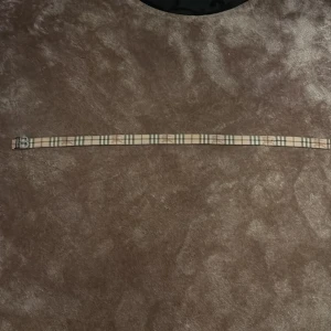 Vändbart bälte med rutigt mönster - Snyggt vändbart bälte med ett klassiskt rutigt mönster på ena sidan och en stilren svart yta på den andra. Bältet har ett silverfärgat spänne format som bokstaven B som står för Burberry