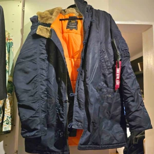 Mörkblå jacka från Alpha Industries - Säljer en mörkblå jacka från Alpha Industries med orange foder och pälsdetaljer på huvan. Jackan har dragkedja och knappar framtill samt en ikonisk 'Remove Before Flight' tag på ärmen. Perfekt för kyliga dagar.
