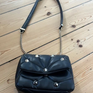 Liten svart axelväska med nitar Zara  - Snygg svart axelväska med kedjerem och coola nitar. Väskan har en quiltad design som ger den en unik look. Använt skick, lite slitage på silver detaljerna, skicka meddelande för mer info och bilder :) 