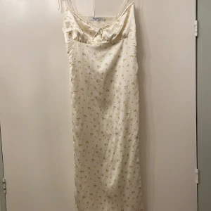 Vit blommig långklänning från na-kd - Säljer en elegant vit långklänning från na-kd med små gula blommor. Klänningen har smala axelband och en subtil slits framtill. Perfekt för sommarens alla tillfällen 🌼