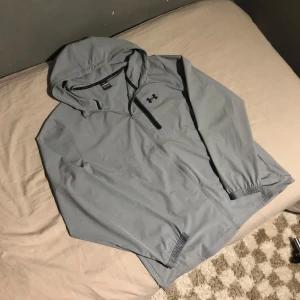 Grå vindjacka från Under Armour - Säljer en grå vindjacka från Under Armour med huva och dragkedja. Jackan har en bröstficka med dragkedja och elastiska ärmslut. Perfekt för blåsiga dagar och sportiga aktiviteter. Några fläckar på bröstet som bör gå bort efter en tvätt (Se på sista bilden)