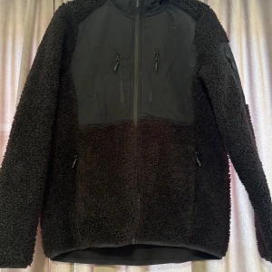 Svart teddyjacka från Five Seasons - Mysig svart teddyjacka från Five Seasons med dragkedja framtill och två praktiska fickor. Perfekt för kyliga dagar med sin fluffiga och varma design. Jackan har långa ärmar och en stilren look.